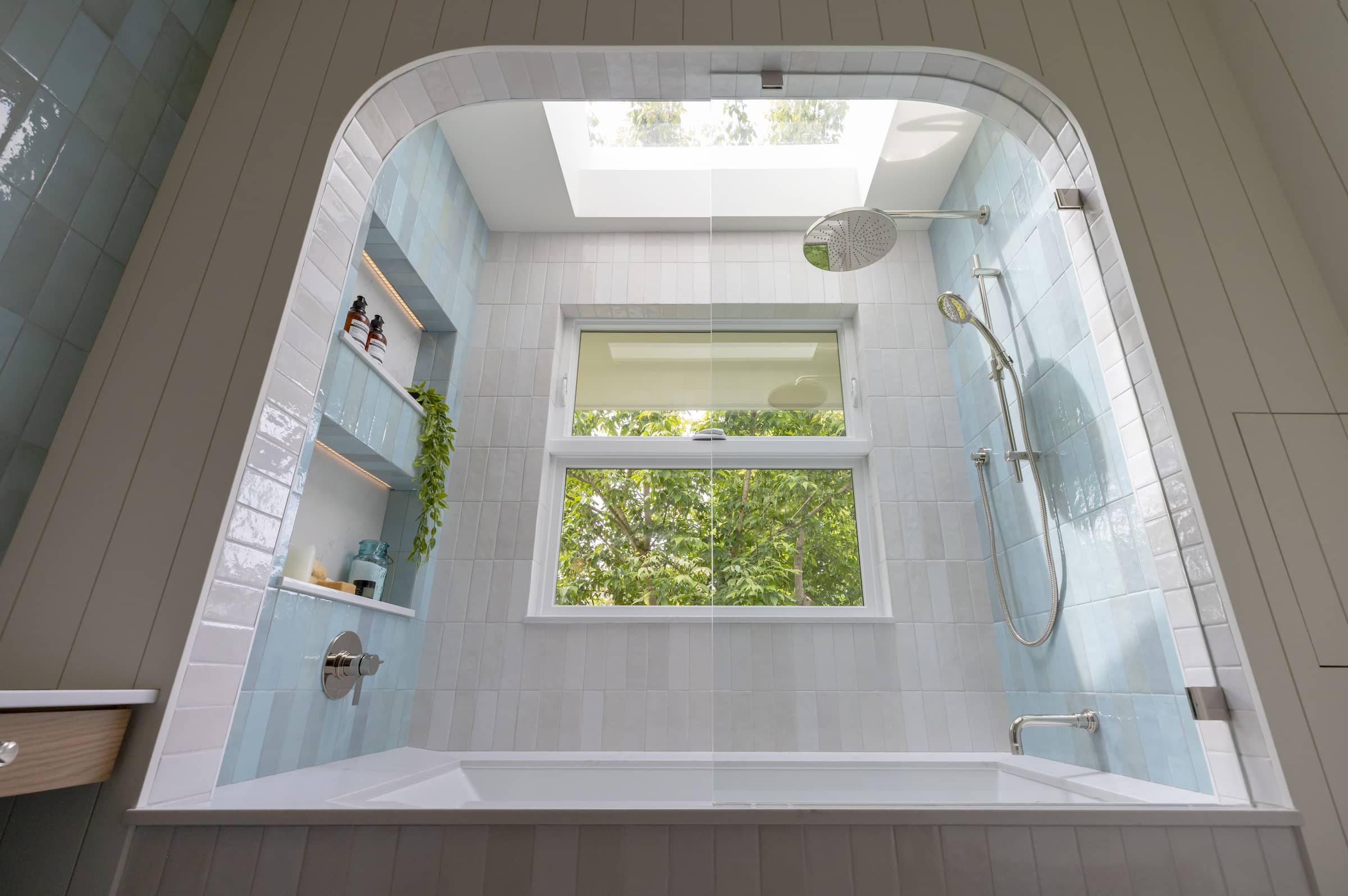 Contemporary bathroom renovation in New Westminster BC featuring an arched tub alcove, skylight, blue shower tile, and large window overlooking a private backyard.